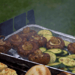 Een verscheidenheid aan groenten zoals grote champignons, gesneden courgette en gegrilde kaas of tofu, bereid in een aluminium bak op de barbecue, met een hand die een champignon omdraait, ideaal om zonder schuldgevoel van te genieten
