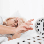 Persoon ligt in bed om 6 uur, reikt naar een witte wekker, ziet er moe of slaperig uit, ter illustratie van het belang van slaap voor een gezonde levensstijl en ochtendroutine.