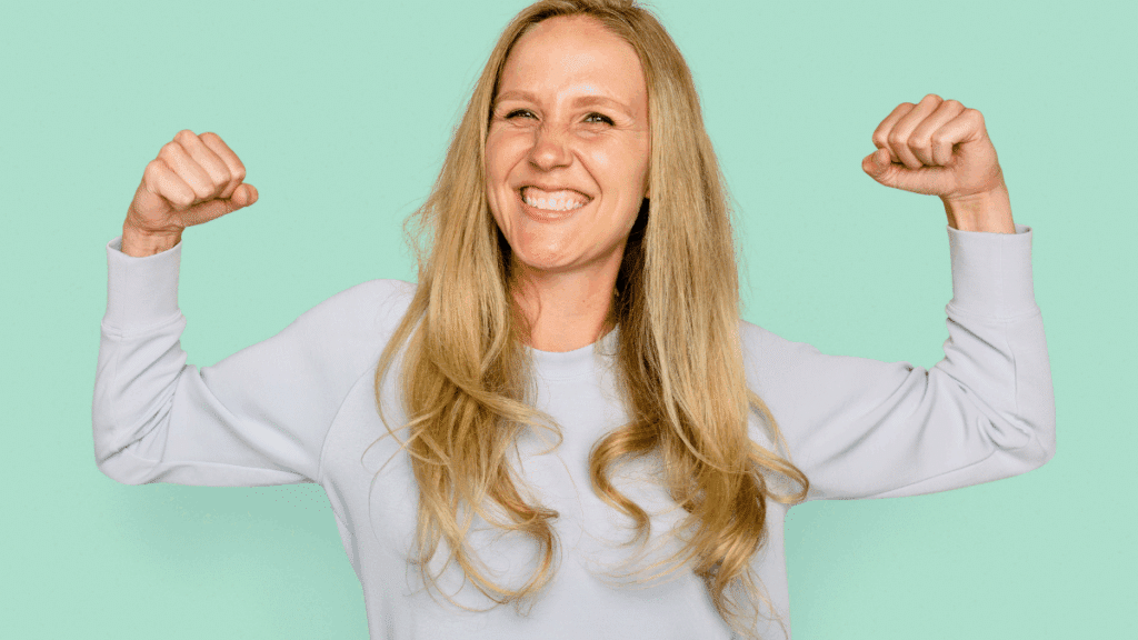 Een blonde vrouw in een lichtblauwe trui toont haar biceps en lacht tegen een lichtblauwe of mintgroene achtergrond, illustratie van hoe gezond voedsel energie geeft en het lichaam sterk maakt