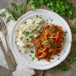 Een bord met Paleo Pulled Pork met koolsalade, geserveerd met een frisse garnering van peterselie. Het gerecht bestaat uit malse, rokerige pulled pork naast een romige koolsalade van gesneden kool en wortelen, perfect voor een gezonde en smaakvolle maaltijd.