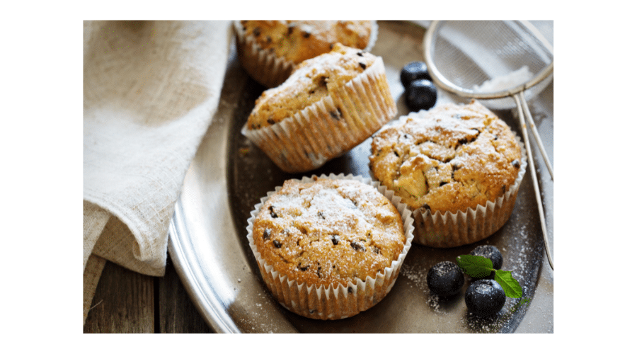 Close up van versgebakken muffins op een metalen schaal, licht bestoven met poedersuiker en omringd door blauwe bessen. De afbeelding straalt een warme en huiselijke sfeer uit en past goed bij paleo bakken en natuurlijke, zelfgemaakte baksels.