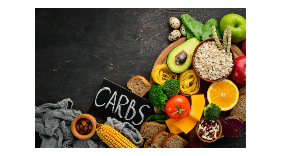 Kleurvolle verzameling koolhydraatrijke en koolhydraatarme voedingsmiddelen op een donkere ondergrond, met onder andere avocado, groenten, fruit, pasta, granen en brood, naast een bordje met het woord Carbs.