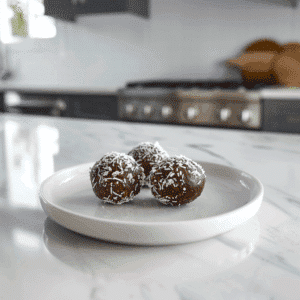 Drie Paleo Cacao Kokos Geluksballetjes, bedekt met geraspte kokos. Gezonde no bake chocoladesnack gemaakt van dadels, noten, cacao en kokos, geschikt als vegan, paleo en suikervrije traktatie.