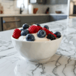 Romige kokosyoghurt met verse bessen, waaronder blauwe bessen, frambozen, bramen en aardbeien, bovenop geserveerd als een kleurrijke en gezonde paleo snack.