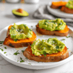 Plakjes geroosterde zoete aardappel belegd met romige geprakte avocado, bestrooid met zeezout, kruiden en zwarte peper. De toast bites hebben een warme oranje kleur en een frisse groene topping.