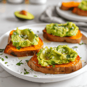 Plakjes geroosterde zoete aardappel belegd met romige geprakte avocado, bestrooid met zeezout, kruiden en zwarte peper. De toast bites hebben een warme oranje kleur en een frisse groene topping.