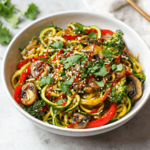 Keto pittige courgettenoedels roerbak met paprika, champignons en broccoli, afgewerkt met sesamzaad en verse koriander in een kom.
