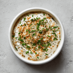 Keto kokos-uien dip in een schaaltje, romig en glad van structuur, gemaakt met kokosroom, kokosmelk en ui, gegarneerd met bieslook, geroosterde kokosvlokken en hennepzaad – een gezonde low-carb dip.