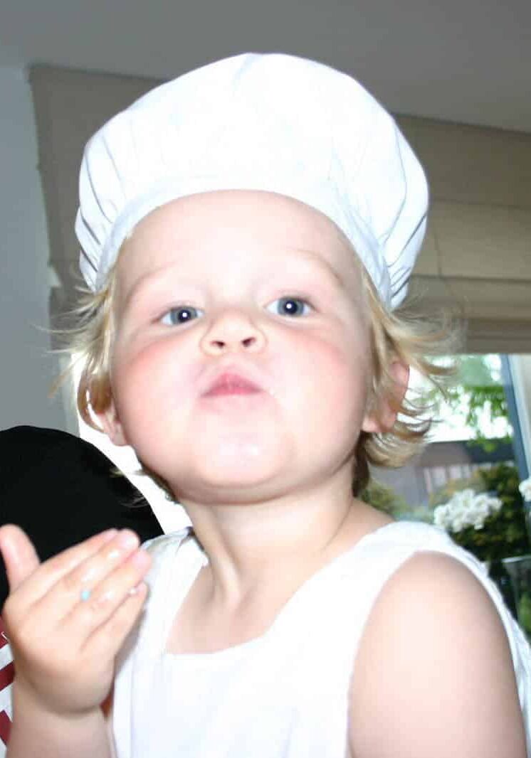 Een jong blond kind met blauwe ogen, gekleed in een witte koksmuts en een mouwloos wit topje, trekt een grappig gezicht met getuite lippen en houdt één hand bij de kin, met een wazige binnenachtergrond — een speels en schattig kookthema-portret.